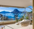 Balcony with large glass windows, view of mountains and a lake, hanging chair, table, lamp