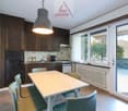 The image shows a modern kitchen and dining area with large windows providing a scenic view. The kitchen features dark wood cabinets, a tiled backsplash, and stainless steel appliances. The dining table is made of light-colored wood and is surrounded by m