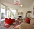 Spacious living room with large windows, red sofa, beige couch, wooden coffee table, shelving unit, and artwork on the wall. The room has access to a balcony or terrace.