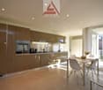 Spacious open-plan kitchen with modern, high-gloss brown cabinets, stainless steel appliances including a dishwasher, and a dining area with a table and chairs next to large windows providing natural light.