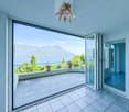 Spacious apartment with large balcony, offering a stunning view of the mountains and lake. The balcony has a tiled floor and is furnished with potted plants. The interior features large windows that provide abundant natural light and access to the balcony