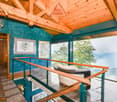 Spacious living room with wooden ceiling beams, teal walls, large windows offering a scenic view, and a cozy lounge area with a rattan chaise.