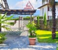 Outdoor patio with a white fence, palm trees, a round table, chairs, and a pergola. The backdrop features a stone wall, a mountain view, and a house.