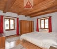 bedroom with wooden bed, wooden wardrobe, wooden floor, two windows with red curtains, white bed sheets, two carpets