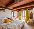 Bedroom with wooden ceiling, bed with floral cover, pillows, desk, chair, windows with yellow curtains, wicker chair, wooden cabinet, painting