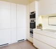 Modern, minimalist kitchen with white cabinets and drawers, built-in appliances including a dishwasher, and a large wall-mounted TV or projection screen.
