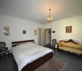 Spacious bedroom with a wooden bed frame, a vintage-style sofa, and a chandelier overhead. The room features white walls, hardwood floors, and various decorative elements such as framed artwork and wall sconces.