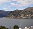 Scenic view of a lakeside town nestled in the hills, with buildings and houses scattered along the slopes. The town is situated on the shore of a large body of water, likely a lake or river. The landscape is dominated by forested hills and mountains in th