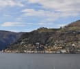 The image shows a scenic view of a town nestled in the hills along a body of water, likely a lake or river. The town features a mix of residential and commercial buildings, with some larger structures visible on the hilltops. The landscape is dominated by