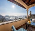 Spacious balcony with ornate wrought-iron railing, offering a panoramic view of the surrounding mountains and a church steeple in the distance. The balcony is furnished with wicker furniture and decorative pillows, creating a cozy and relaxing outdoor spa