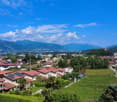 The image shows a residential area with a mix of single-family homes and multi-unit buildings. The houses have red tile roofs and are surrounded by lush greenery, including trees and gardens. The area is situated in a valley, with mountains visible in the