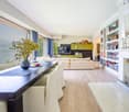 Spacious living room with large windows offering a scenic view, hardwood flooring, a long dining table, a comfortable sofa set, a TV unit with shelves, and a chandelier overhead.