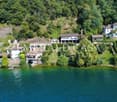 Multiple houses on a hillside overlooking a lake, stonework and brick exterior, green water, surrounded by trees and plants, a small boat in the foreground