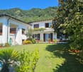Two-story house, white exterior, awnings, green plants, red flowers in planters, swimming pool, expansive garden, mountainous background