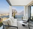 Balcony with a couch, center table, two armchairs, sliding glass door, railings, panoramic view of a lake and cityscape.