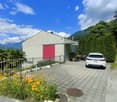 2-story building with a gray exterior, a red garage door, and surrounded by lush greenery and flowers. There is a paved driveway with a parked car in front of the building.
