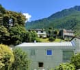 Two-story house with a green exterior, surrounded by lush greenery and mountains in the background. The house has a balcony or terrace visible.