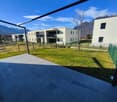 The image shows a modern apartment complex with several multi-story buildings surrounded by a well-maintained grassy area. The buildings have a minimalist, contemporary design with large windows and balconies. The complex appears to be situated in a sceni