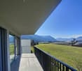 A balcony with a metal railing overlooks a wide open field and mountain range