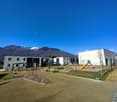 The image shows a residential complex with several multi-story buildings, a playground with swings and slides, and a grassy area with benches. The complex is surrounded by mountains in the background, creating a scenic setting.