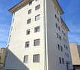 Modern white high-rise building, large windows with shutters, landscaped garden in front, multiple floors with several apartments.