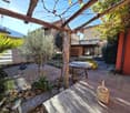 Small gravel garden with plants and trees, wooden pergola, table and basket, and distant buildings