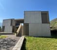 Modern concrete house, raised ground floor, stairs leading to entrance, large windows, flat roof