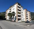 A multi-story residential building with balconies, located on a street with a parking area and a signboard. The building has a modern design with a combination of white and brown colors. There are palm trees and a fence in front of the building, and a mou