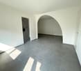 An empty room with gray tiled flooring, white walls, and a small archway on the left side leading to another room. An oven and cabinets are visible in the room through the archway. A power outlet is seen on the wall on the right.