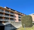 A multi-story residential building with multiple balconies, surrounded by greenery and parking spaces, set against a backdrop of mountains.