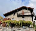 Stone house, brown roof, wooden balcony, raised ground floor, red leafy plants