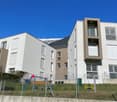 Modern apartment building with white and brown exterior, multiple windows, balconies, and a fenced playground area.