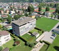 Aerial view of a residential building with a parking lot, surrounded by other buildings, trees, and a green field.