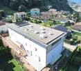 Aerial view of a modern white building with solar panels on the roof, surrounded by other houses and greenery.