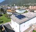 Aerial view of a white building with solar panels on the roof, surrounded by other buildings and trees, with a mountain in the background.
