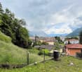 hillside garden with grass, trees, and a fence, overlooking a town with buildings and mountains