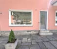 Pink building with a window and a door, a small plant in a pot, a paved entrance, a small balcony above the door, a small staircase leading to the entrance.