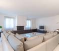 living room, white walls, large grey sofa, glass coffee table, two plants, windows with white curtains, white tiles, television