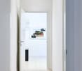 Open door leading to hallway, hardwood flooring, clean white walls, closed door at end of hallway