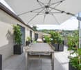 Terrace with wooden table and chairs, potted plants, parasol, and a view of the cityscape.