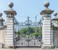 Grand old entrance gate with stone pillars, wrought iron gates, small wall-mounted lamps, bicycle riders at the entrance.