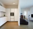 Modern, minimalist kitchen with white cabinets, stainless steel appliances including a dishwasher, and a sleek countertop. The kitchen opens up to a living room area with a black sofa and a TV stand.