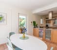 Open-concept kitchen and dining area with white walls, hardwood floors, and large windows overlooking a balcony or terrace. The kitchen features wooden cabinets, a granite countertop, and a built-in dishwasher. The dining table has a white top and mint gr