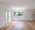 Spacious living room with large windows providing a view of the outdoor greenery, hardwood flooring, and a sliding glass door leading to a balcony or terrace.