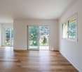 Spacious living room with large windows offering a scenic view of the surrounding landscape and buildings. The room features hardwood flooring and white walls, creating a bright and airy atmosphere.