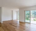 Spacious living room with large windows offering a scenic view of the surrounding landscape, including a church steeple and buildings. The room has hardwood flooring and white walls, creating a bright and airy atmosphere.