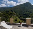 Terrace with lounge chair, tiled floor, metal railing, potted plant, distant mountain landscape with greenery and clouds.