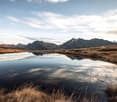a pond with grass on both sides, mountains in the background, clouds in the sky