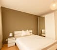 Spacious bedroom with beige walls, white bed with pillows, nightstand with lamp, and a crystal chandelier overhead. There appears to be a balcony or terrace visible through the window.