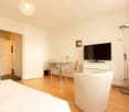 Spacious bedroom with hardwood floors, white walls, a large TV mounted on the wall, a small dining table with two chairs, and a white sofa. The room is well-lit with a crystal chandelier overhead.